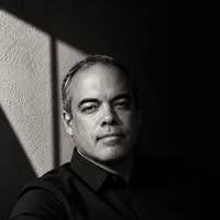 Black-and-white portrait of Mark Dalton in a black shirt, lit by hard window light against a textured wall